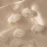 Light Ivory Organza Flower Earrings A pair of dangle earrings with layered white organza flower petals hanging from thin silver wires, displayed on a draped beige satin fabric background.