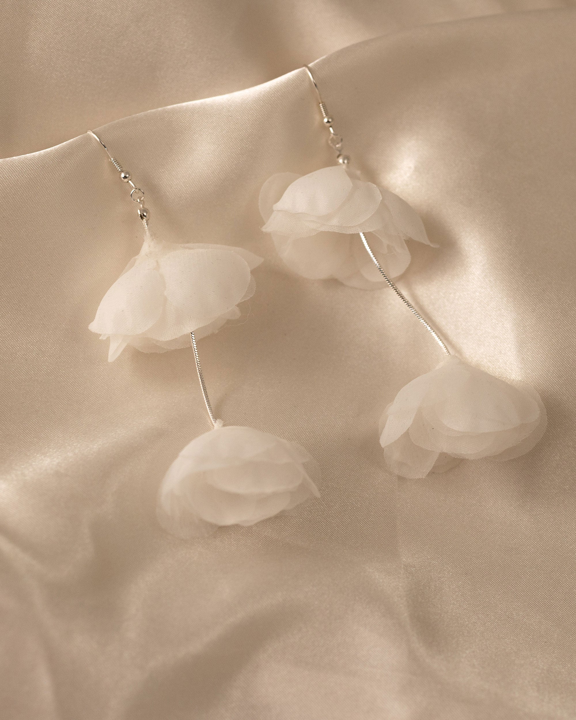Light Ivory Organza Flower Earrings A pair of dangle earrings with layered white organza flower petals hanging from thin silver wires, displayed on a draped beige satin fabric background.