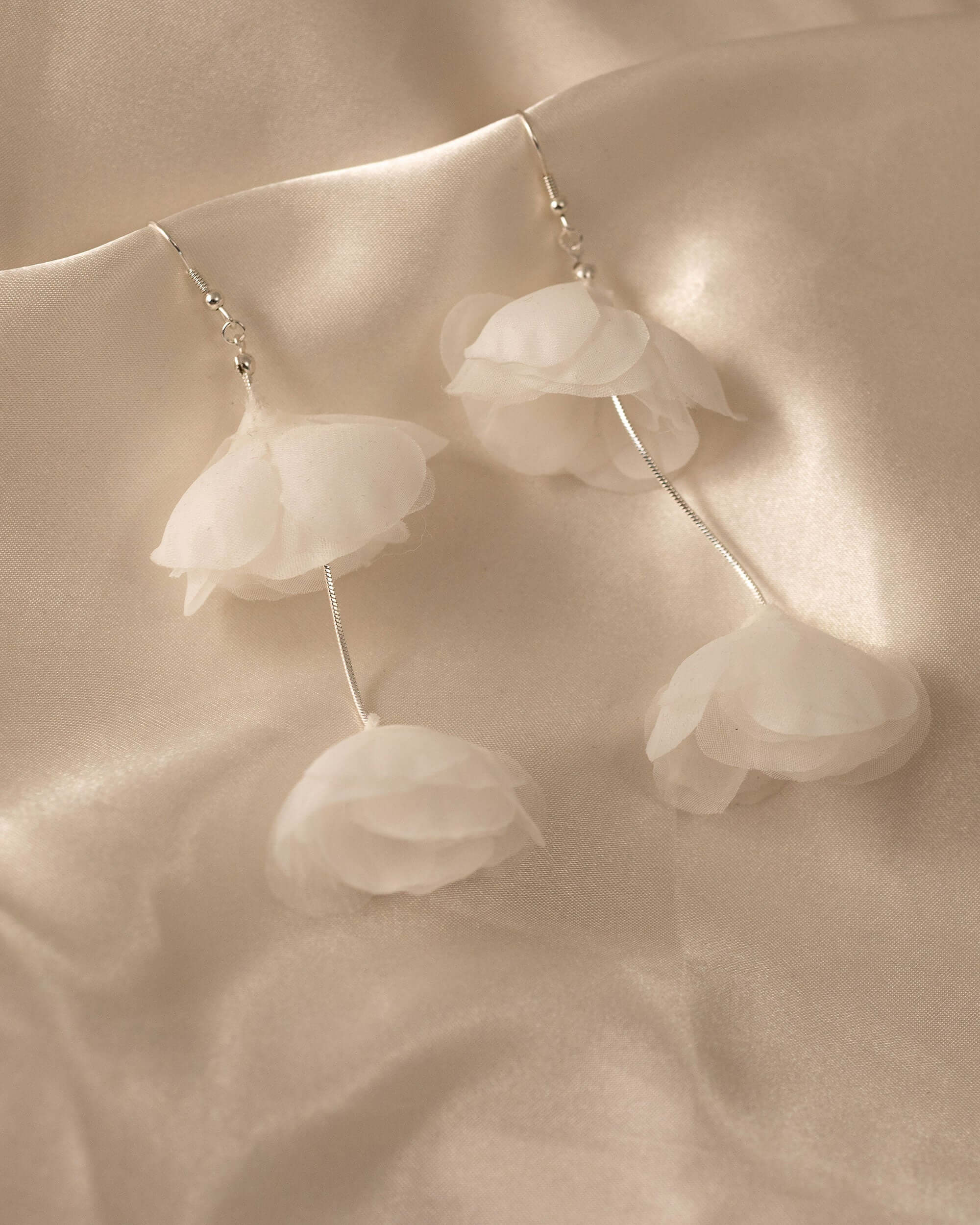 Light Ivory Organza Flower Earrings A pair of dangle earrings with layered white organza flower petals hanging from thin silver wires, displayed on a draped beige satin fabric background.