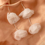Light Ivory Organza Flower Earrings A pair of dangling flower earrings made of layered translucent pale pink fabric petals, suspended from thin silver wires on a peach satin fabric background.