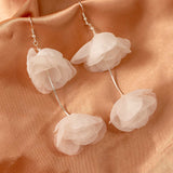 Light Ivory Organza Flower Earrings A pair of dangling flower earrings made of layered translucent pale pink fabric petals, suspended from thin silver wires on a peach satin fabric background.