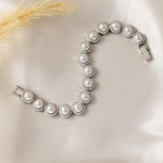 Tennis Style Pearl CZ Bracelet A silver-toned pearl bracelet with round off-white pearls and halo settings, arranged in an S-shape on a cream satin fabric background with a dried grass stem prop.