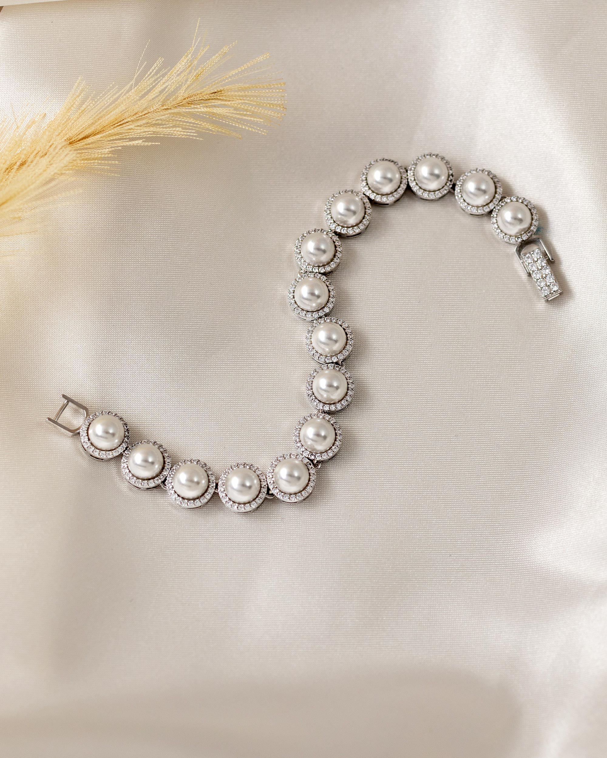 Tennis Style Pearl CZ Bracelet A silver-toned pearl bracelet with round off-white pearls and halo settings, arranged in an S-shape on a cream satin fabric background with a dried grass stem prop.