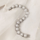Tennis Style Pearl CZ Bracelet A silver-toned bracelet featuring a series of round off-white faux pearls surrounded by small clear crystals, displayed on a soft white tulle fabric background.