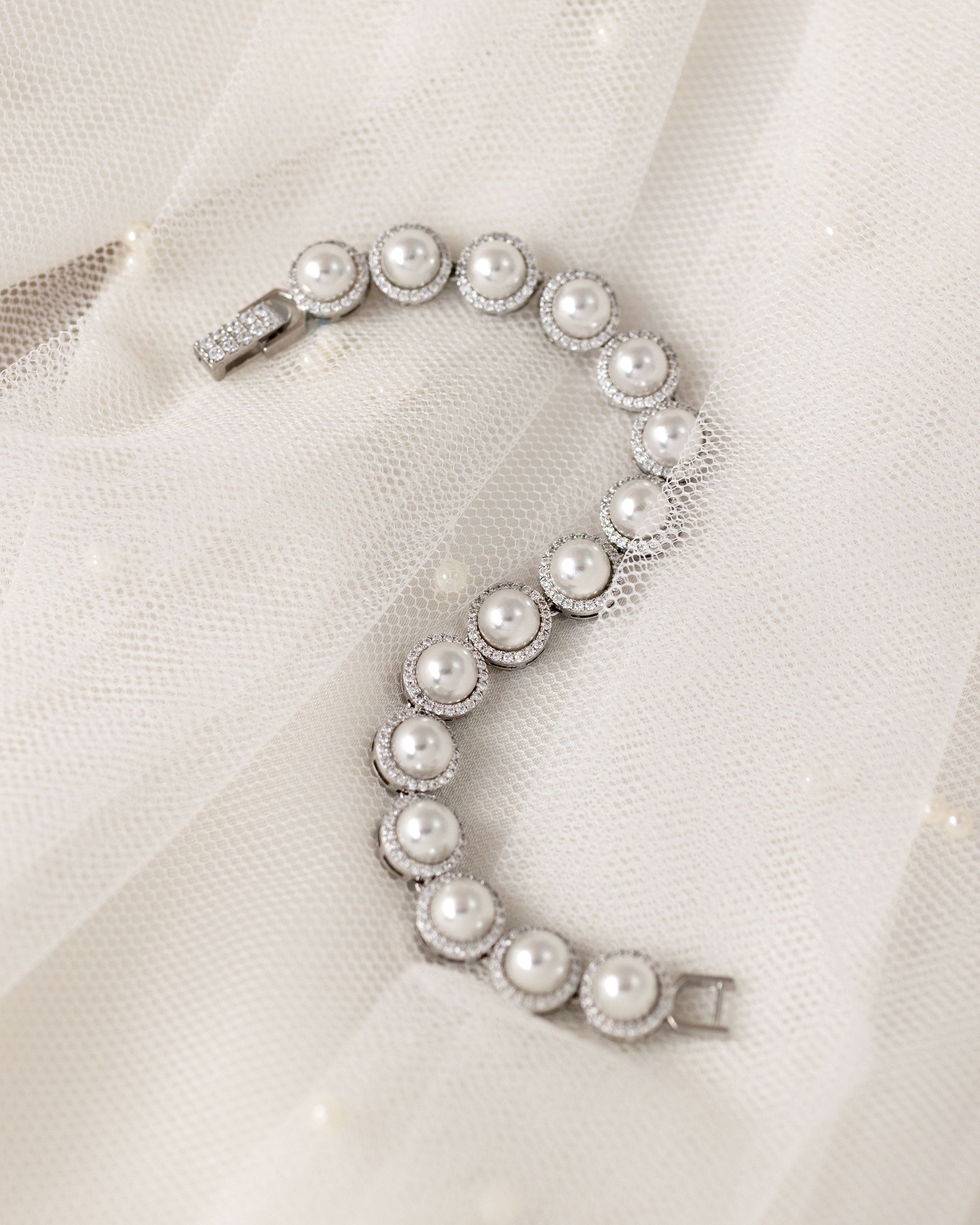 Tennis Style Pearl CZ Bracelet A silver-toned bracelet featuring a series of round off-white faux pearls surrounded by small clear crystals, displayed on a soft white tulle fabric background.