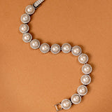 Tennis Style Pearl CZ Bracelet A pearl and diamond bracelet arranged in a curved S-shape on a warm brown background. The bracelet features large off-white pearls surrounded by small clear diamonds in silver-toned settings, with a clasp visible at the top and bottom.