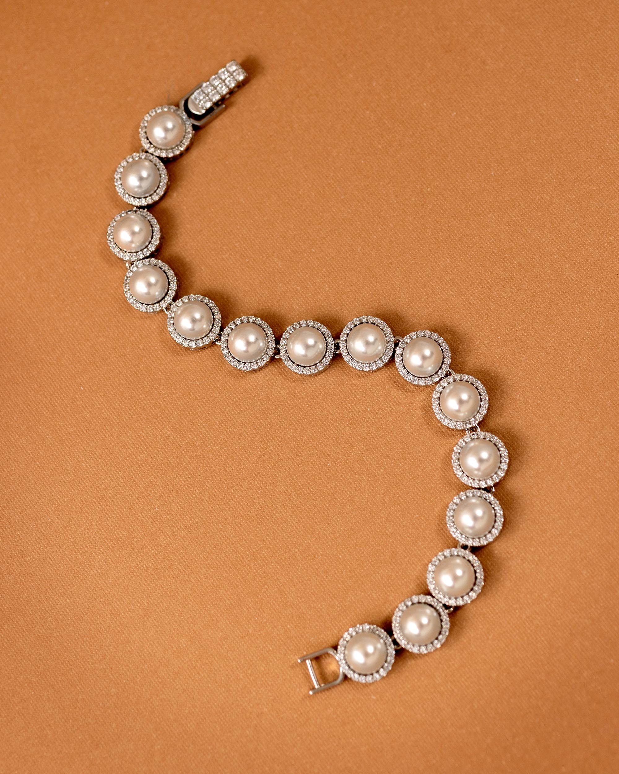 Tennis Style Pearl CZ Bracelet A pearl and diamond bracelet arranged in a curved S-shape on a warm brown background. The bracelet features large off-white pearls surrounded by small clear diamonds in silver-toned settings, with a clasp visible at the top and bottom.