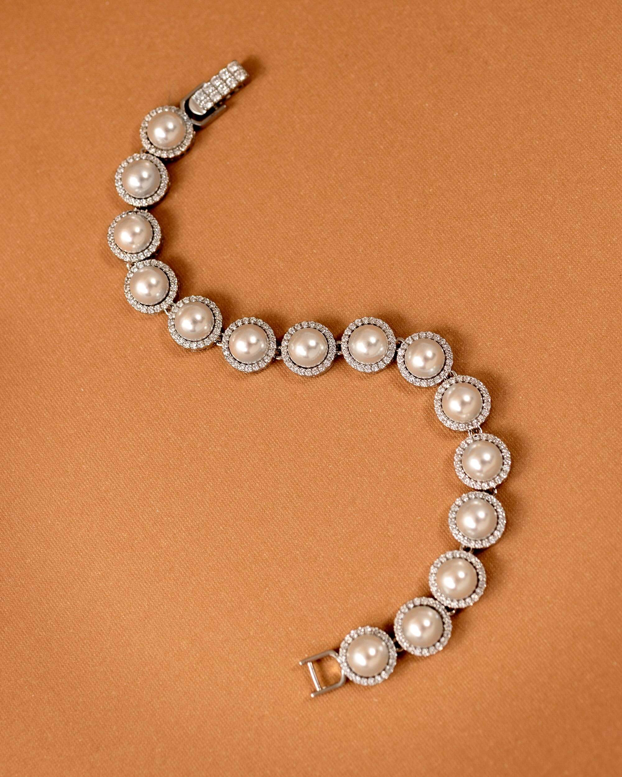 Tennis Style Pearl CZ Bracelet A pearl and diamond bracelet arranged in a curved S-shape on a warm brown background. The bracelet features large off-white pearls surrounded by small clear diamonds in silver-toned settings, with a clasp visible at the top and bottom.