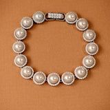 Tennis Style Pearl CZ Bracelet A pearl and crystal bracelet arranged in a circle on a textured brown background. The bracelet features large off-white pearls surrounded by small clear crystals, with a silver-toned clasp at the top.