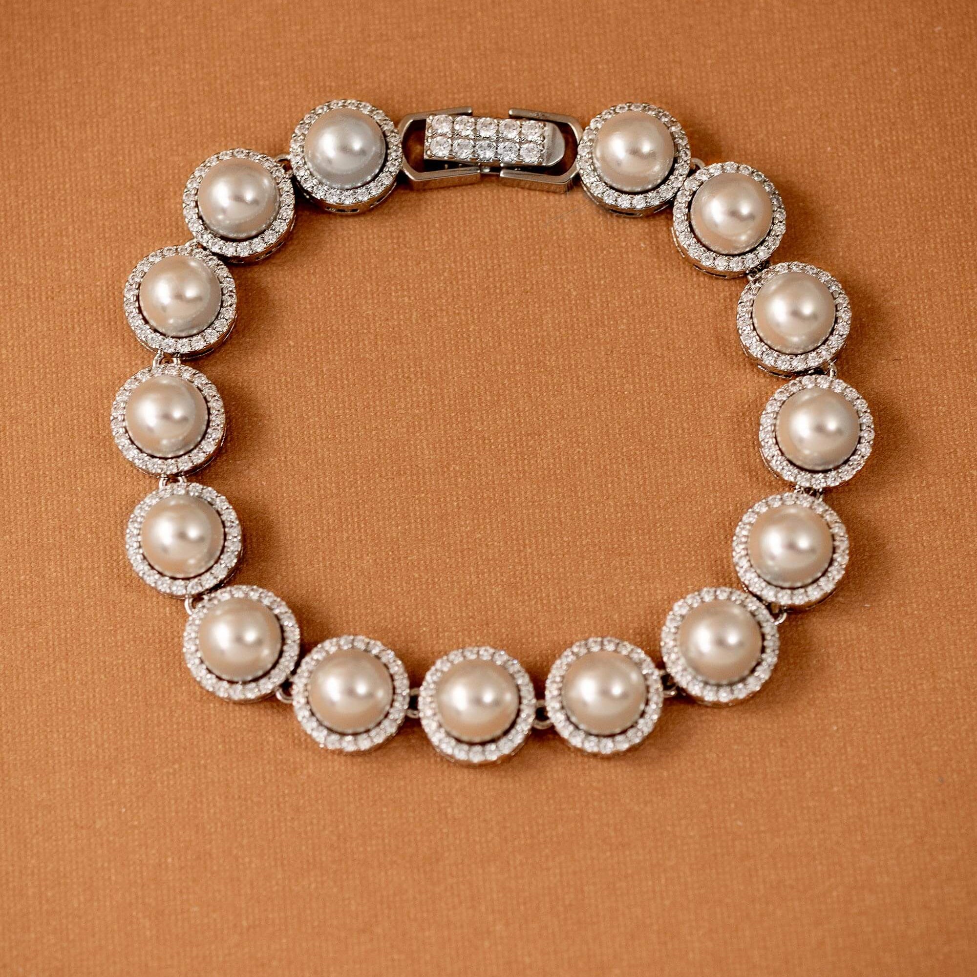 Tennis Style Pearl CZ Bracelet A pearl and crystal bracelet arranged in a circle on a textured brown background. The bracelet features large off-white pearls surrounded by small clear crystals, with a silver-toned clasp at the top.