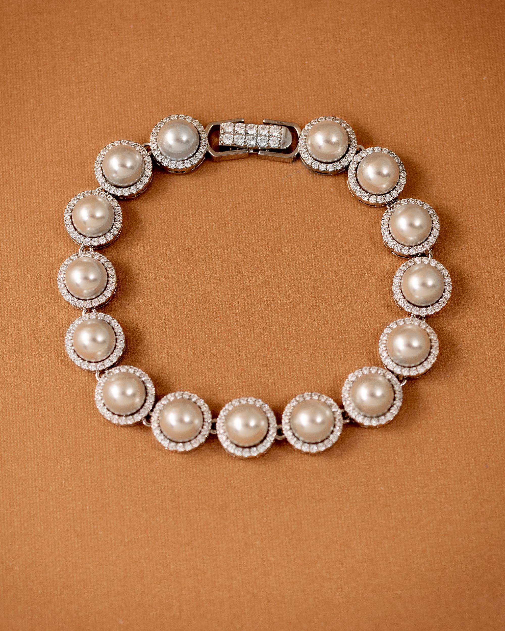 Tennis Style Pearl CZ Bracelet A pearl and crystal bracelet arranged in a circle on a textured brown background. The bracelet features large off-white pearls surrounded by small clear crystals, with a silver-toned clasp at the top.