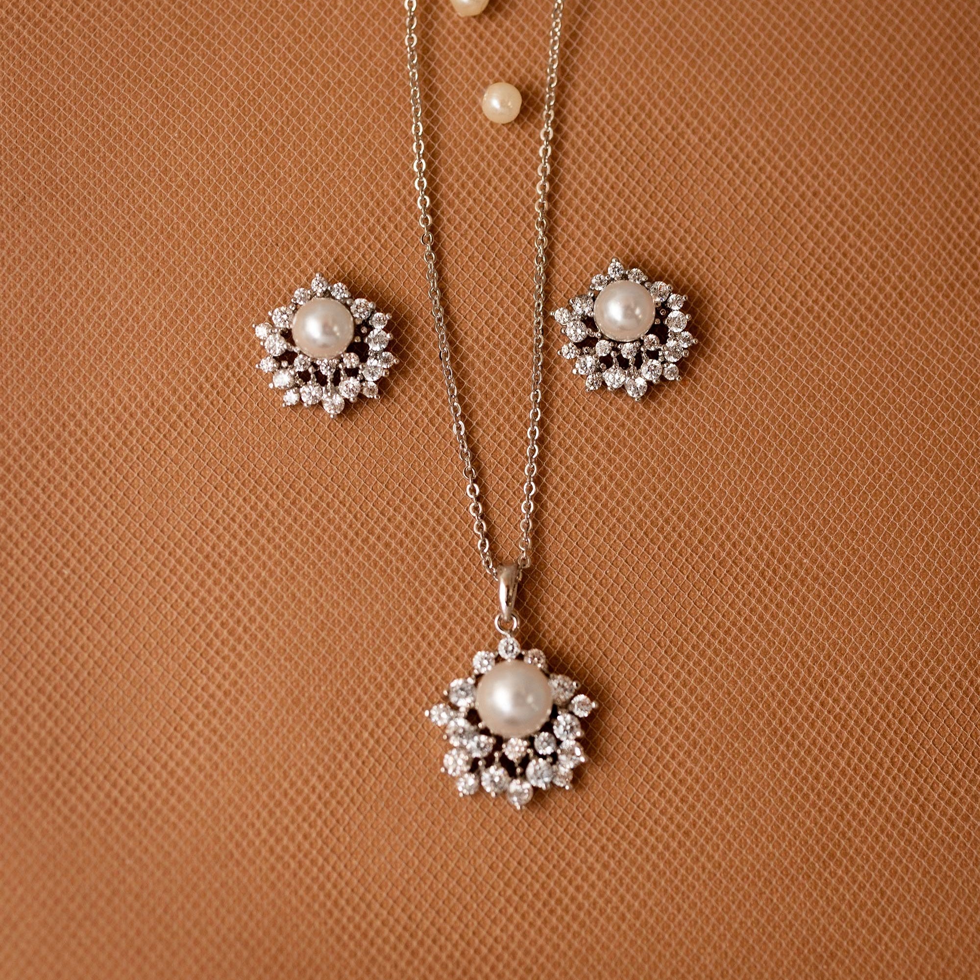 A silver-tone jewelry set featuring a pendant necklace and matching earrings, each with a central white pearl surrounded by clear stones, displayed on a textured brown background.