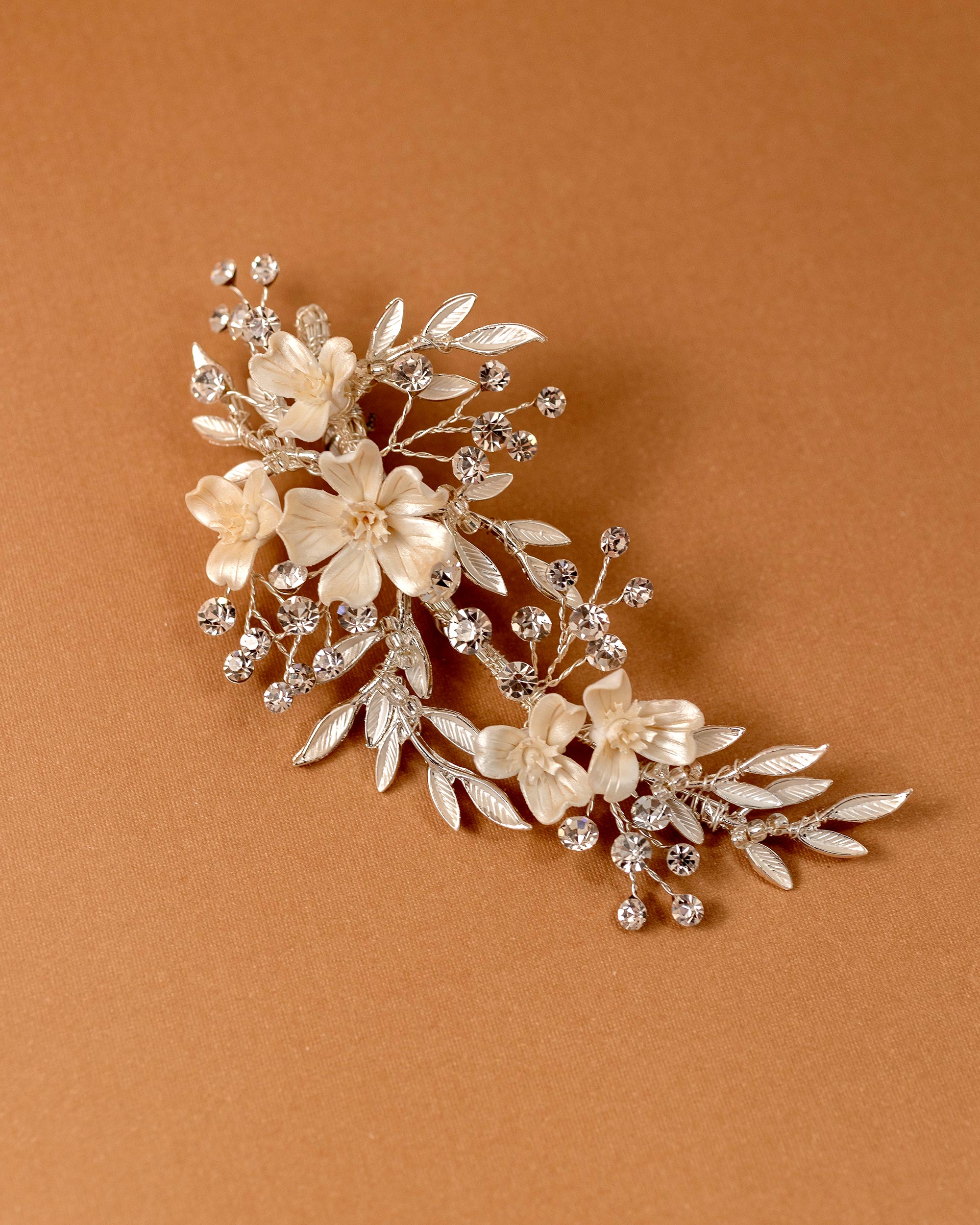 Porcelain Ivory Flowers and Crystals Bridal Clip A floral bridal hair claw clip featuring cream-colored faux flowers and silver-toned leaves, accented with clear rhinestones, displayed on a warm brown background.