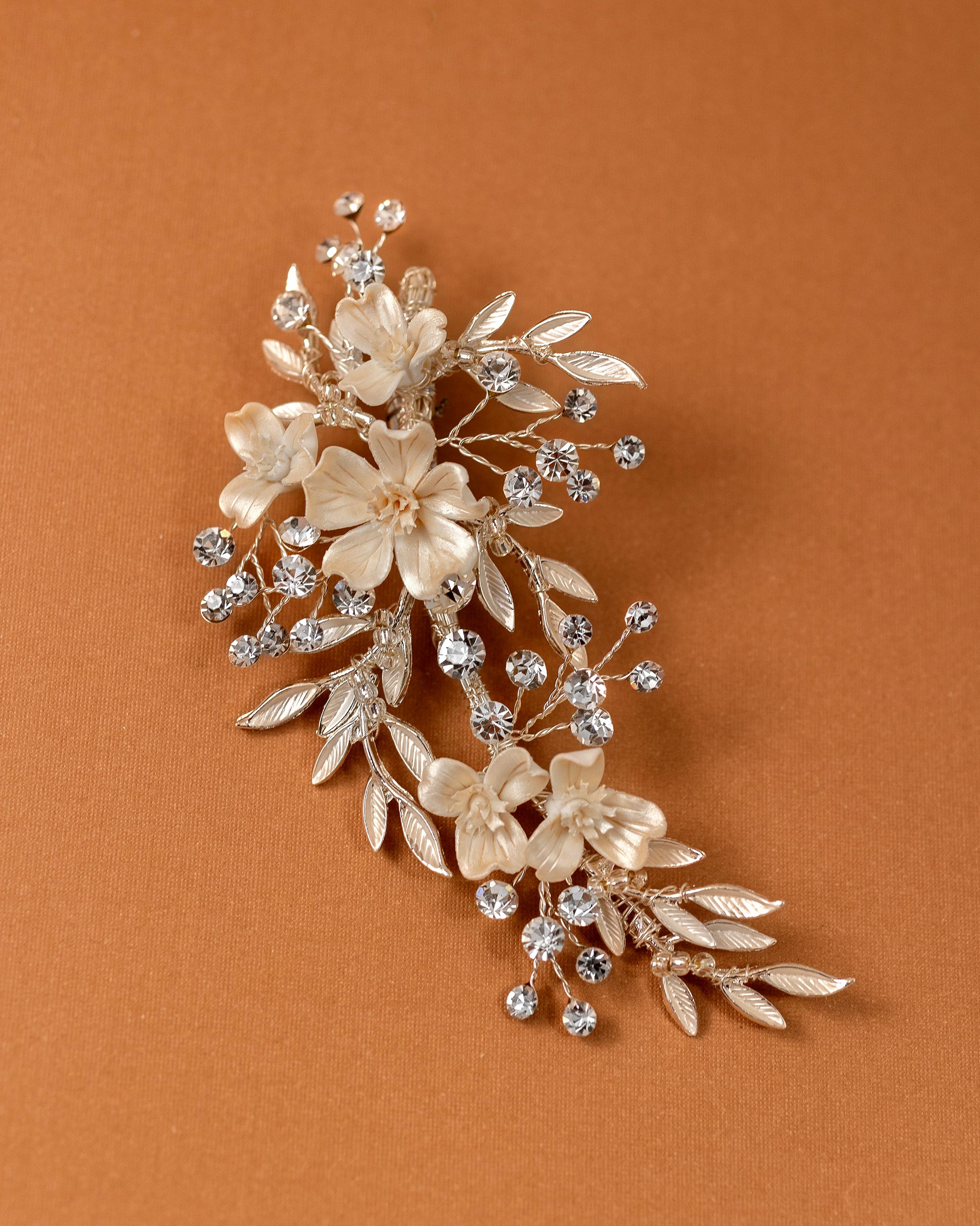 Porcelain Ivory Flowers and Crystals Bridal Clip A floral and leaf hair accessory brooch featuring cream-colored faux flowers, silver-toned leaves, and clear rhinestones, displayed on a warm brown background.