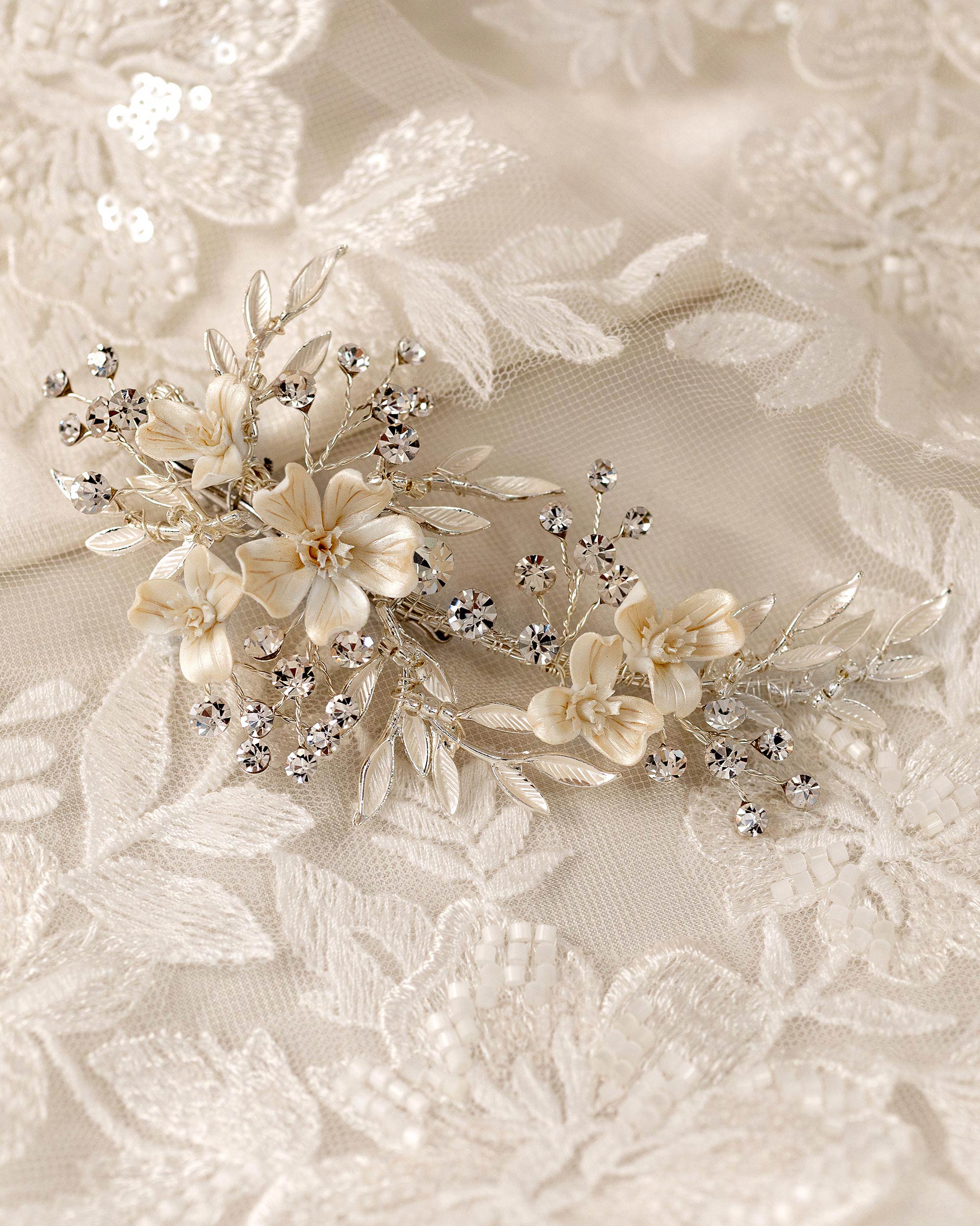 Porcelain Ivory Flowers and Crystals Bridal Clip A delicate bridal hairpiece featuring cream satin flowers, small crystal beads, and silver-toned leaves, displayed on a matching cream lace fabric background.