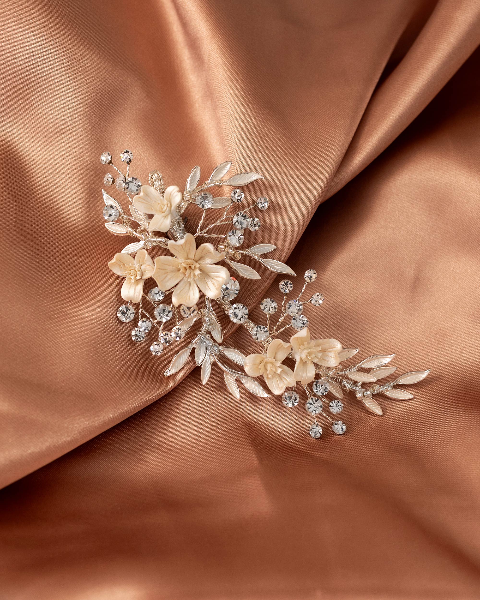 Porcelain Ivory Flowers and Crystals Bridal Clip A floral bridal hair accessory featuring cream and champagne embroidered flowers, silver-toned leaves, and clear rhinestones, displayed on draped champagne satin fabric.