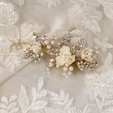 Pearled Porcelain Flower Hair Clip A delicate bridal hairpiece featuring cream-colored fabric flowers, faux pearls, and gold-toned crystal-embellished leaves, displayed on a white lace fabric background.