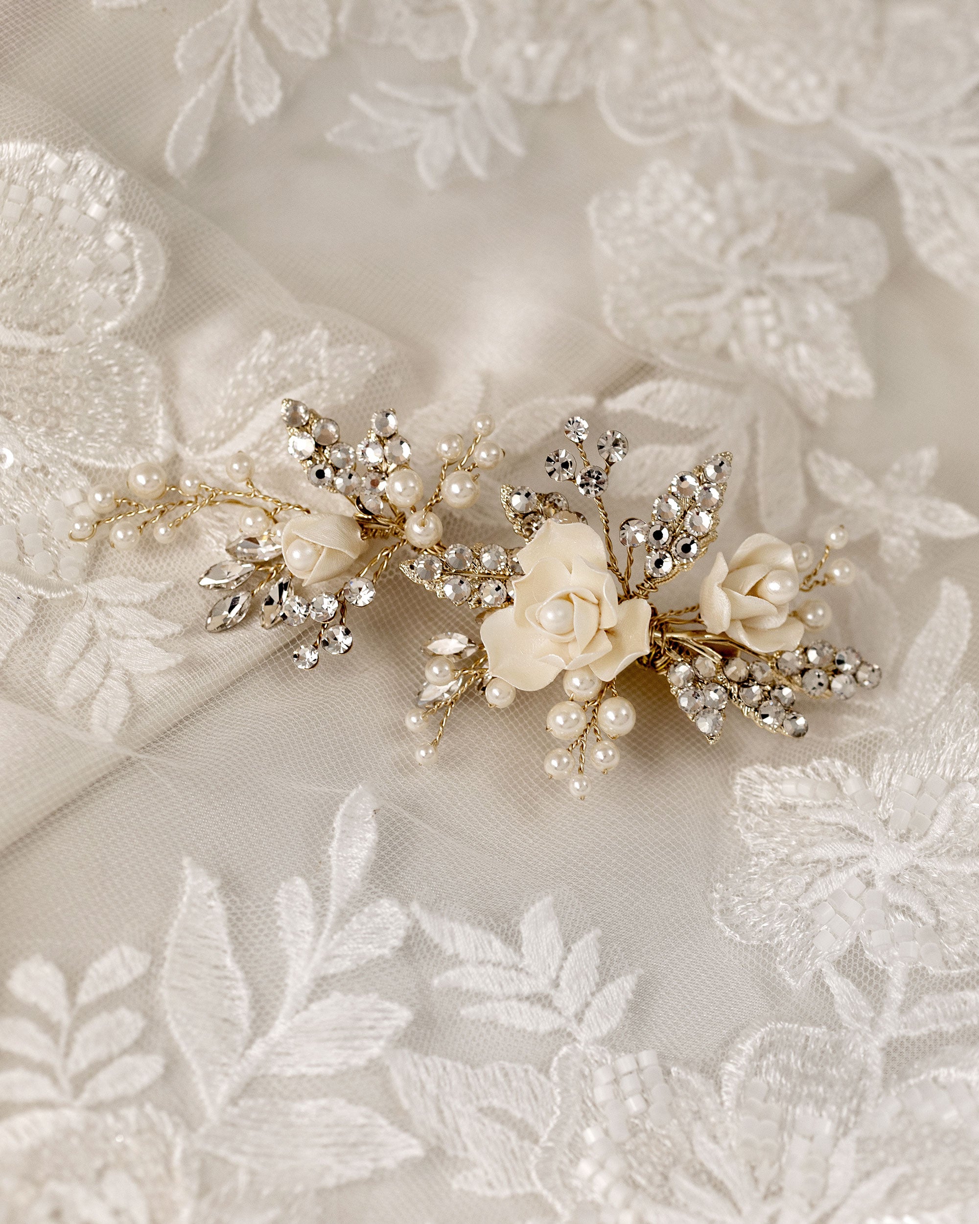 Pearled Porcelain Flower Hair Clip A delicate bridal hairpiece featuring cream-colored fabric flowers, faux pearls, and gold-toned crystal-embellished leaves, displayed on a white lace fabric background.