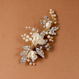 Pearled Porcelain Flower Hair Clip A gold-toned floral hair accessory featuring cream-colored faux pearls and clear rhinestones, arranged in a cascading floral design on a solid brown background.