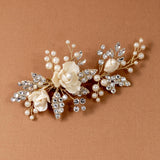 Pearled Porcelain Flower Hair Clip A gold-toned floral hair claw clip decorated with cream-colored faux roses, pearl-like beads, and clear rhinestones, displayed on a textured brown background.