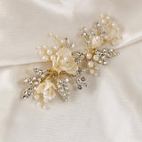 Pearled Porcelain Flower Hair Clip A gold bridal hairpin brooch featuring cream-colored faux roses, white pearls, and clear rhinestones, displayed on a draped cream satin fabric.