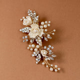 Pearled Porcelain Flower Hair Clip A gold-toned floral hair pin featuring cream-colored faux pearls and clear rhinestones, displayed on a solid warm brown background.