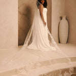 Royal Wedding Veil with Organza Flowers Cassandra Lynne A woman wearing a strapless ivory wedding gown with a long sheer tulle train and floral lace trim, standing in a warm beige architectural setting with a large vase in the background.