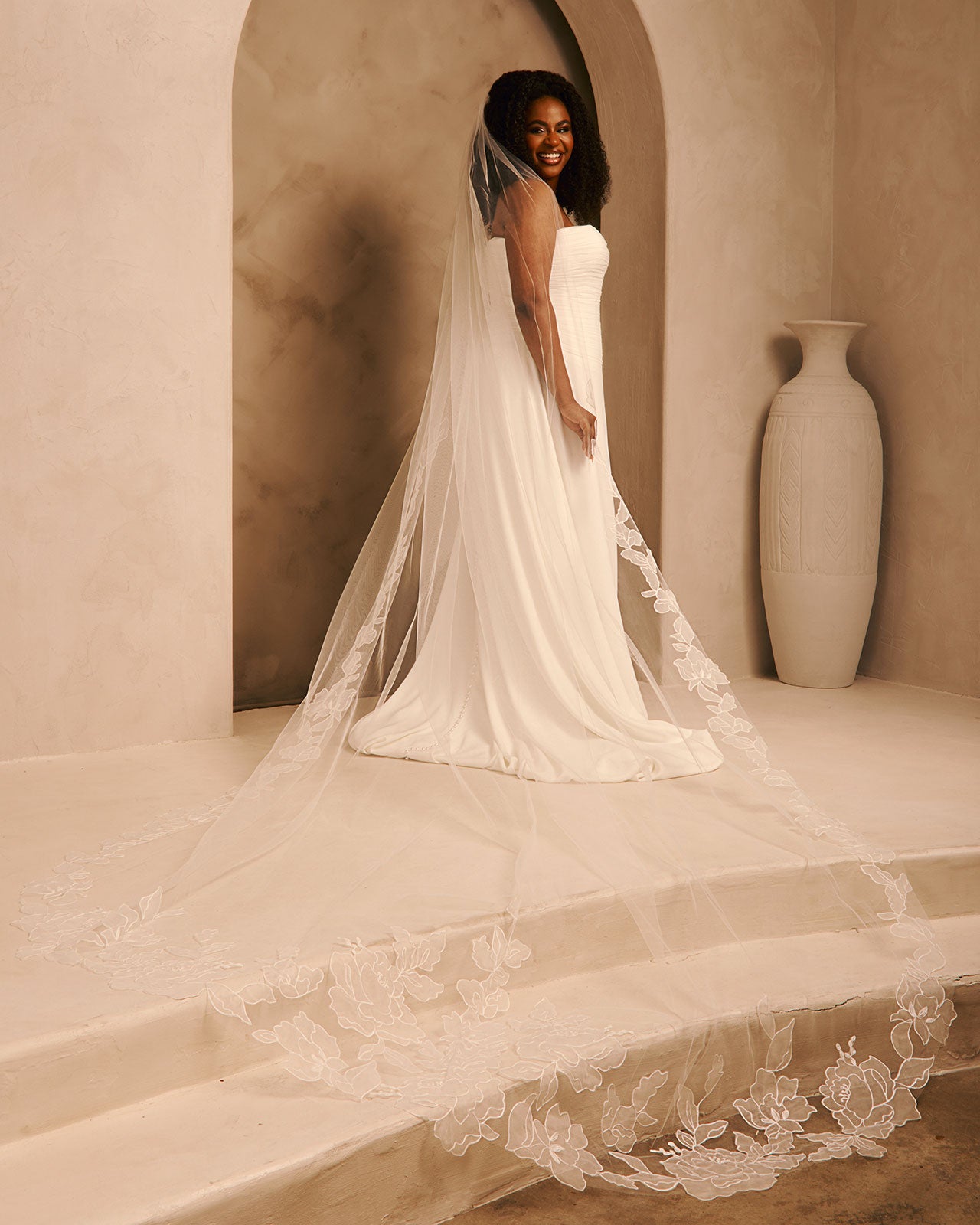 Royal Wedding Veil with Organza Flowers Cassandra Lynne A woman wearing a strapless ivory wedding gown with a long sheer tulle train and floral lace trim, standing in a warm beige architectural setting with a large vase in the background.