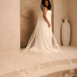 Royal Wedding Veil with Organza Flowers Cassandra Lynne - A woman wearing a strapless ivory wedding gown with a long sheer veil and floral lace trim, standing in a softly lit architectural setting with beige walls, an archway, and a large vase. 