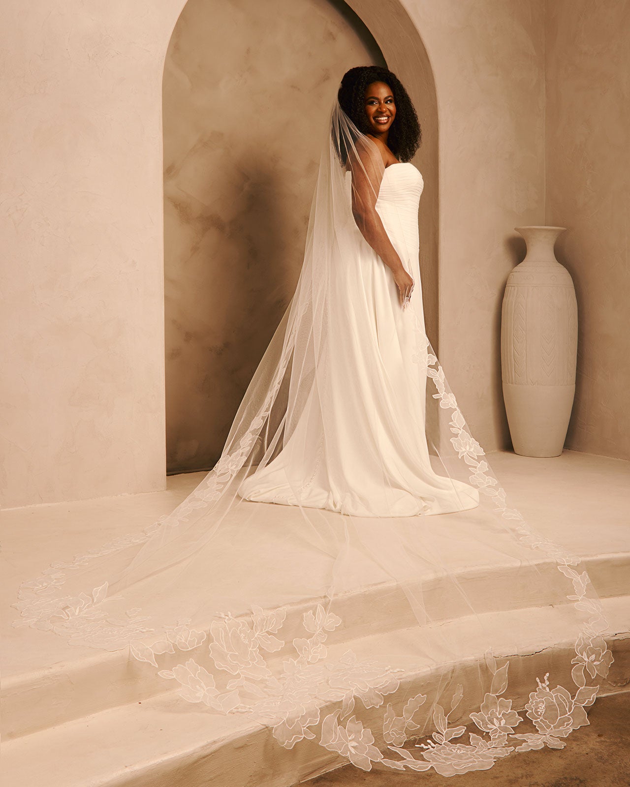 Royal Wedding Veil with Organza Flowers Cassandra Lynne - A woman wearing a strapless ivory wedding gown with a long sheer veil and floral lace trim, standing in a softly lit architectural setting with beige walls, an archway, and a large vase. 