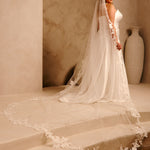 Royal Cathedral Veil with Flower Appliques and Sequins Cassandra Lynne -A woman wearing a wedding gown and long sheer veil with floral lace trim, standing on stone steps in a warm beige interior with a large vase in the background.