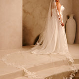 Royal Cathedral Veil with Flower Appliques and Sequins Cassandra Lynne -A woman wearing a wedding gown and long sheer veil with floral lace trim, standing on stone steps in a warm beige interior with a large vase in the background.