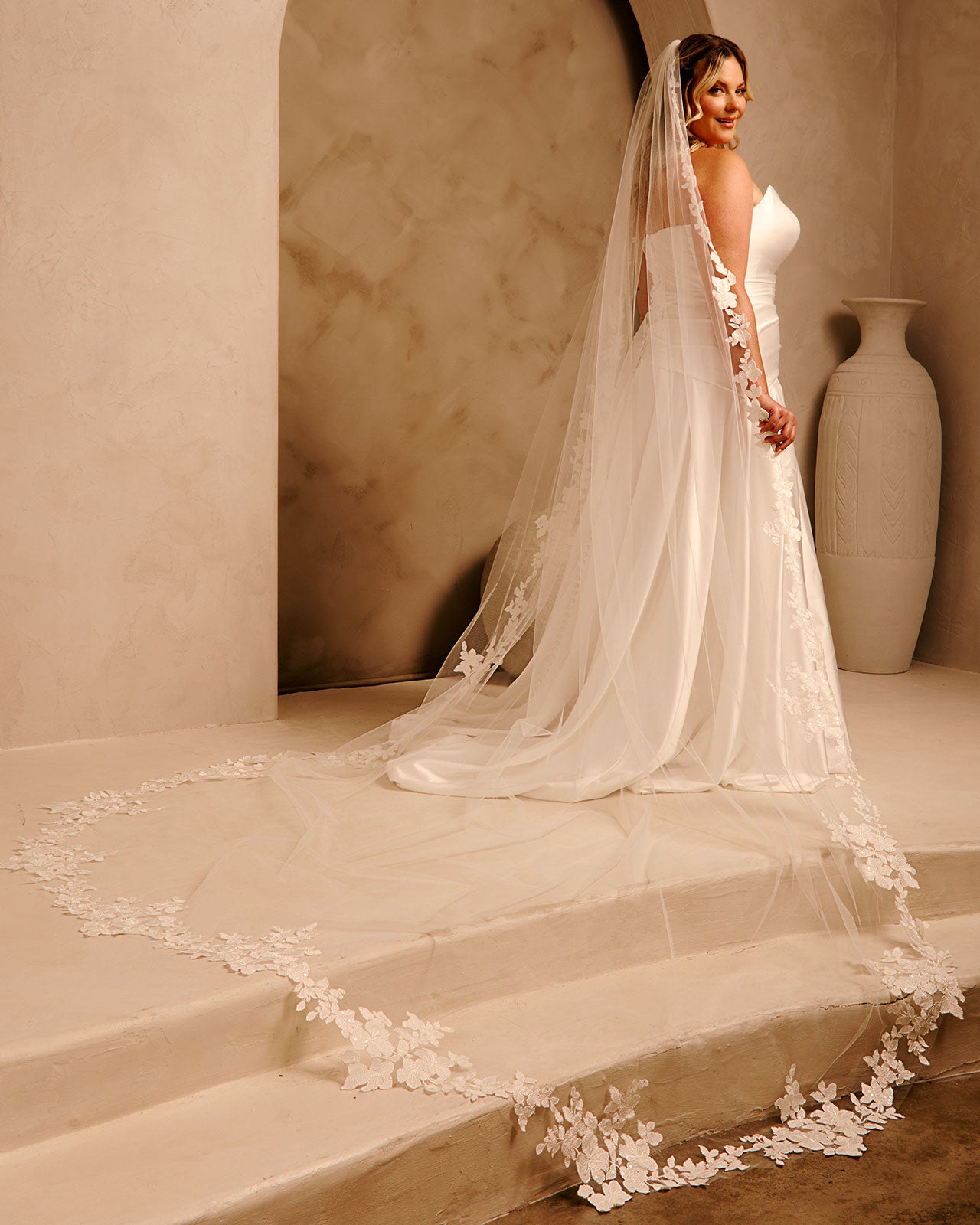 Royal Cathedral Veil with Flower Appliques and Sequins Cassandra Lynne -A woman wearing a wedding gown and long sheer veil with floral lace trim, standing on stone steps in a warm beige interior with a large vase in the background.