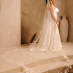 Royal Cathedral Veil with Flower Appliques and Sequins Cassandra Lynne - A woman wearing a wedding gown and long sheer veil with floral lace trim stands on stone steps in a warm beige interior with an arched wall and a large vase in the background.