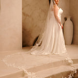 Royal Cathedral Veil with Flower Appliques and Sequins Cassandra Lynne - A woman wearing a wedding gown and long sheer veil with floral lace trim stands on stone steps in a warm beige interior with an arched wall and a large vase in the background.