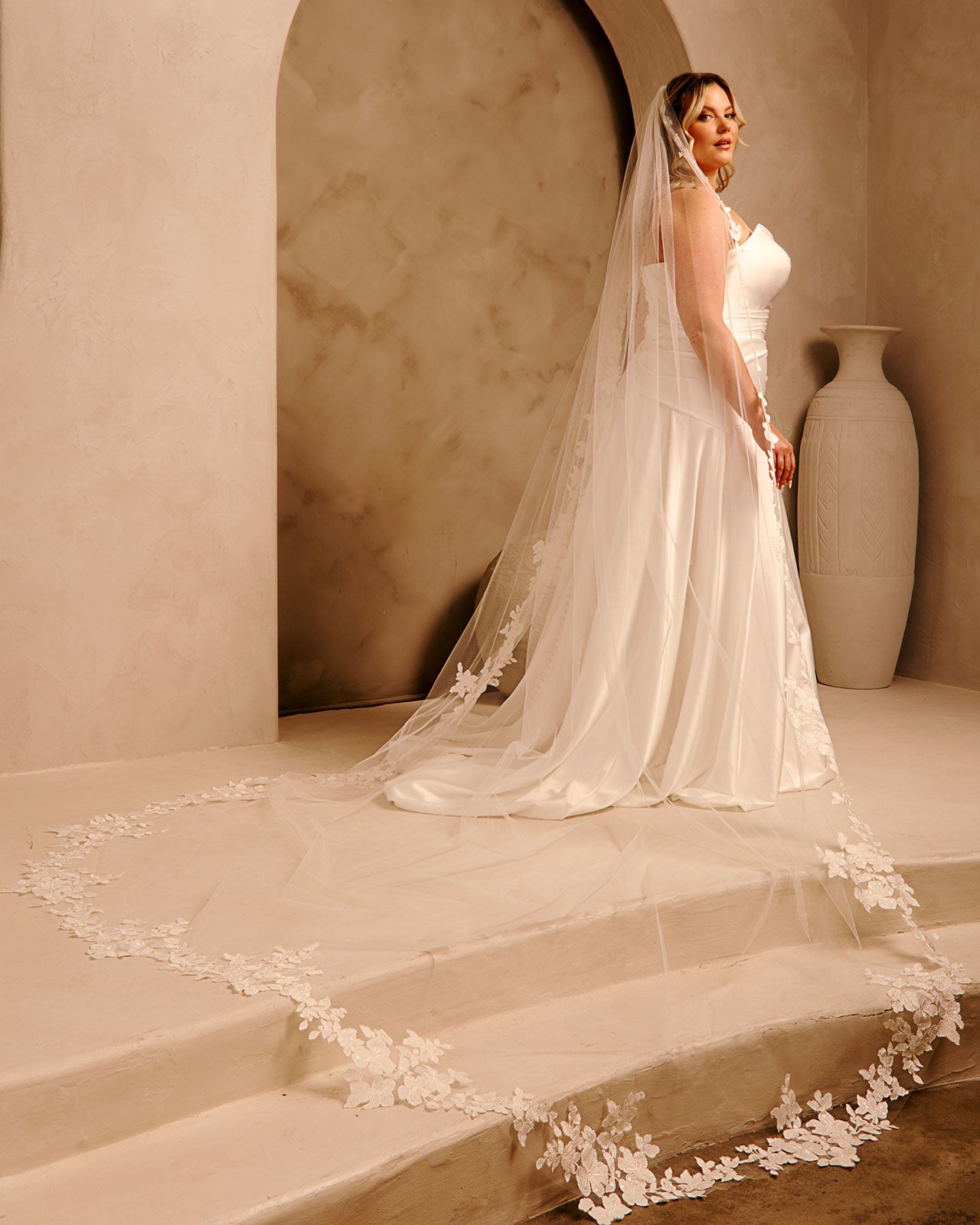Royal Cathedral Veil with Flower Appliques and Sequins Cassandra Lynne - A woman wearing a wedding gown and long sheer veil with floral lace trim stands on stone steps in a warm beige interior with an arched wall and a large vase in the background.