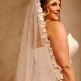 Royal Cathedral Veil with Flower Appliques and Sequins Cassandra Lynne -A woman in a ivory wedding dress and long sheer veil with floral lace trim, photographed against a warm beige studio background.