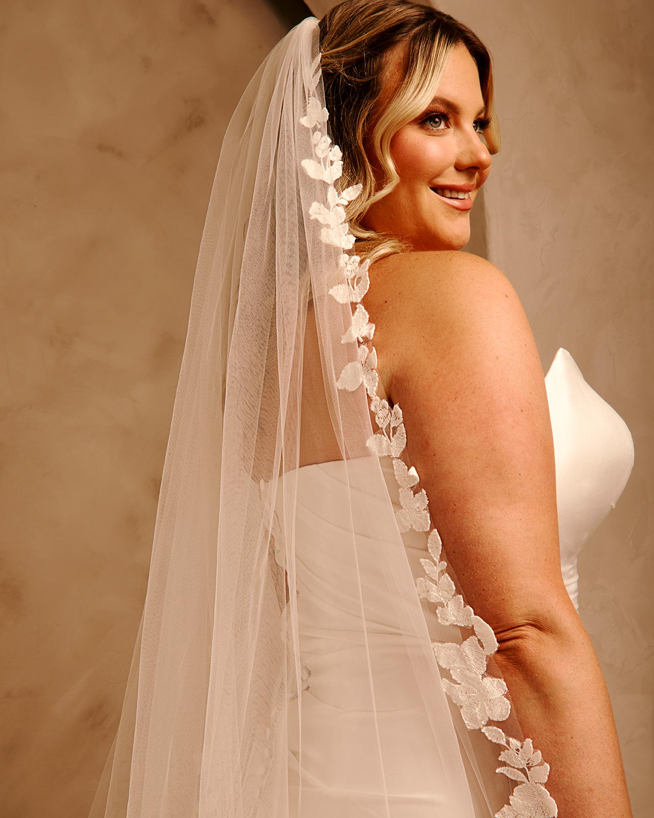 Royal Cathedral Veil with Flower Appliques and Sequins Cassandra Lynne -A woman in a ivory wedding dress and long sheer veil with floral lace trim, photographed against a warm beige studio background.
