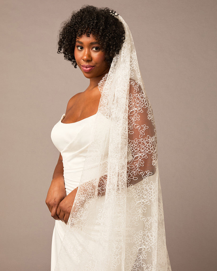 Woman wearing a white wedding dress with a long lace veil against a brown background All over lace veil Cassandra Lynne