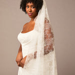 Woman wearing a white wedding dress with a long lace veil against a brown background All over lace veil Cassandra Lynne