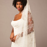 Woman wearing a white wedding dress with a long lace veil against a brown background All over lace veil Cassandra Lynne
