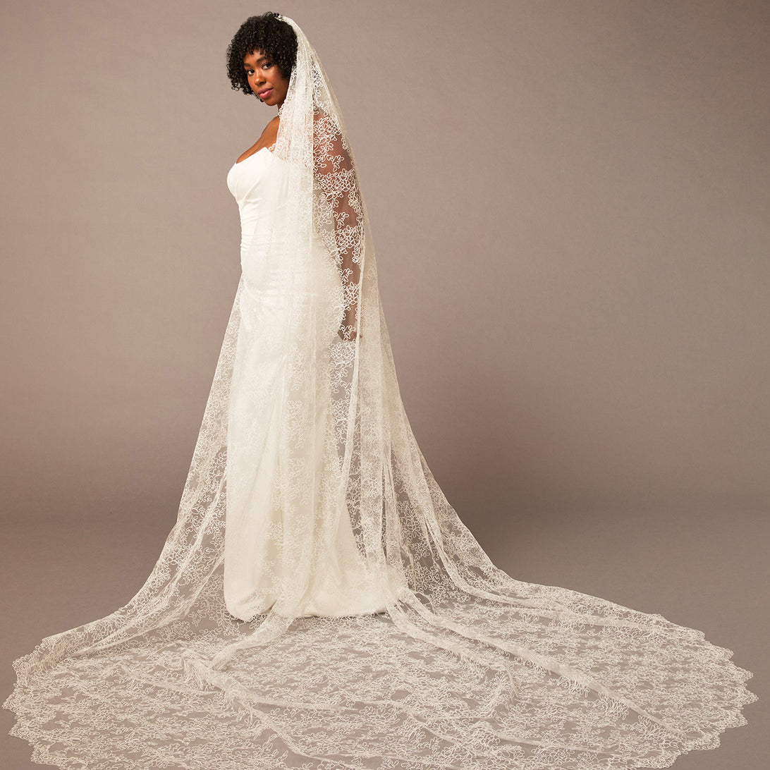 Woman wearing a white wedding dress with a long lace veil against a brown background All over lace veil Cassandra Lynne