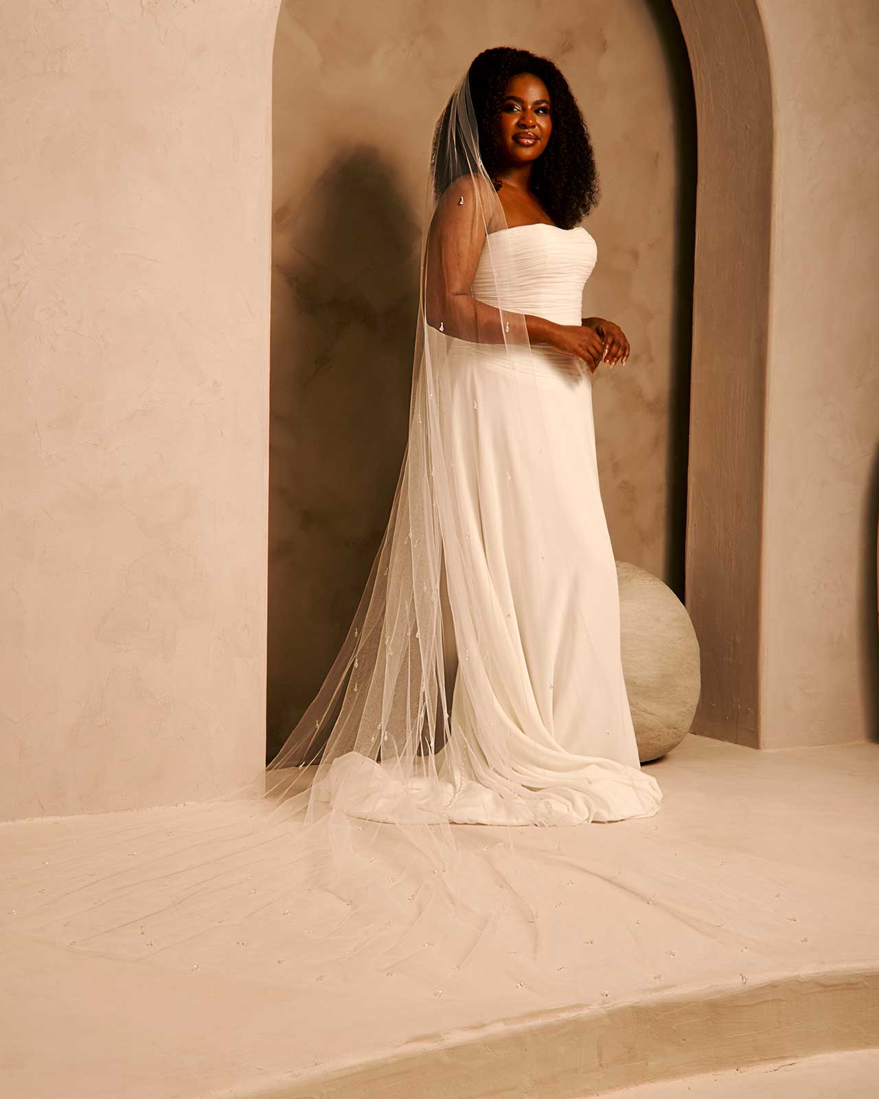 Bridal Veil with Crystal Raindrop Scatter A woman wearing a strapless ivory wedding gown with a long sheer veil stands in a softly lit architectural setting with beige walls and arches.