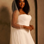 Bridal Veil with Crystal Raindrop Scatter cassandra Lynne - A woman wearing a strapless ivory wedding gown with a long sheer veil, standing against a warm beige textured wall.