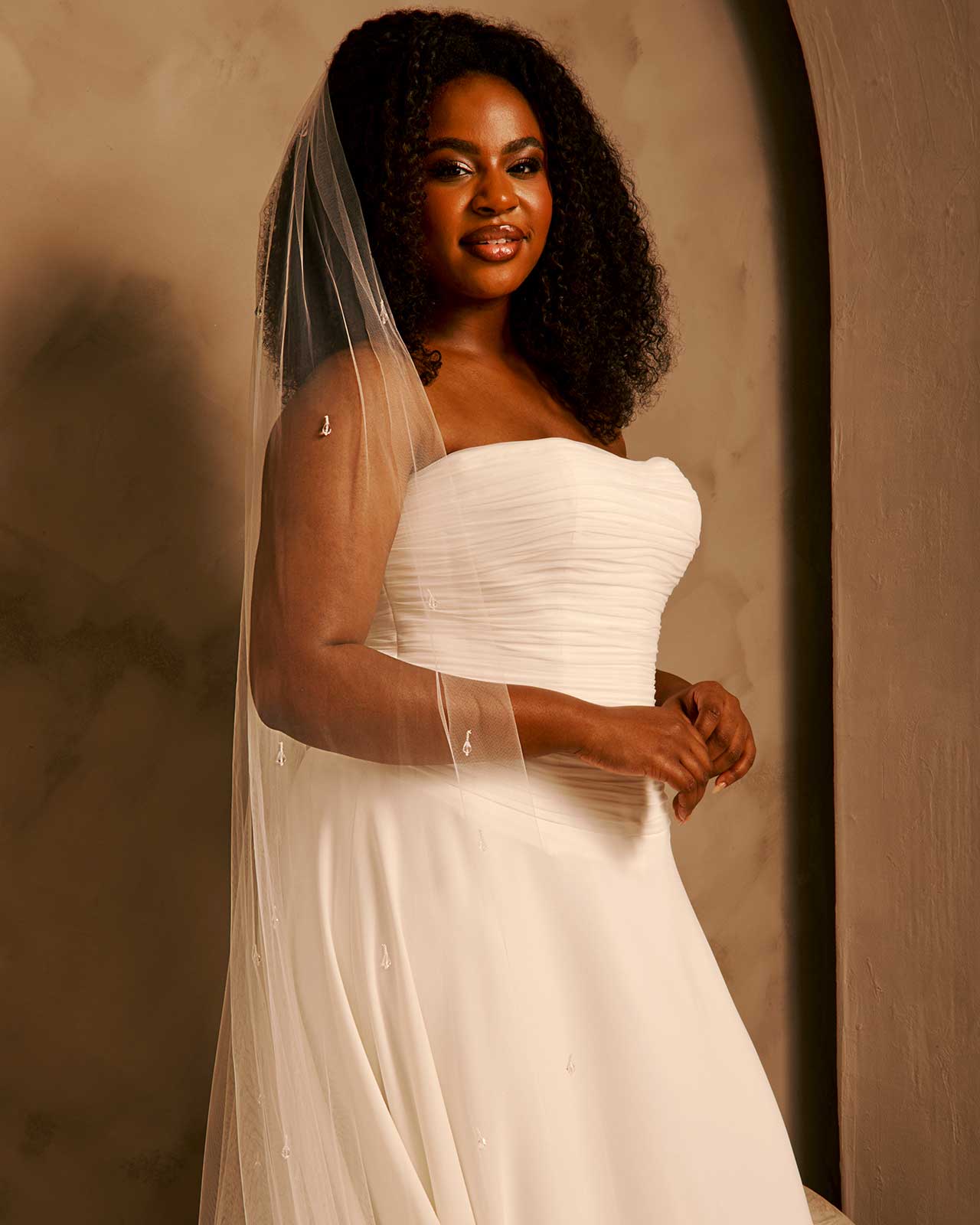 Bridal Veil with Crystal Raindrop Scatter cassandra Lynne - A woman wearing a strapless ivory wedding gown with a long sheer veil, standing against a warm beige textured wall.