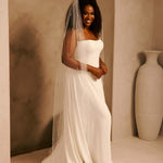 Bridal Veil with Crystal Raindrop Scatter A woman wearing a strapless ivory wedding gown and a long sheer veil, standing indoors against a warm beige arched wall with a large white vase to the right.