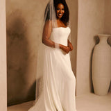 Bridal Veil with Crystal Raindrop Scatter A woman wearing a strapless ivory wedding gown and a long sheer veil, standing indoors against a warm beige arched wall with a large white vase to the right.