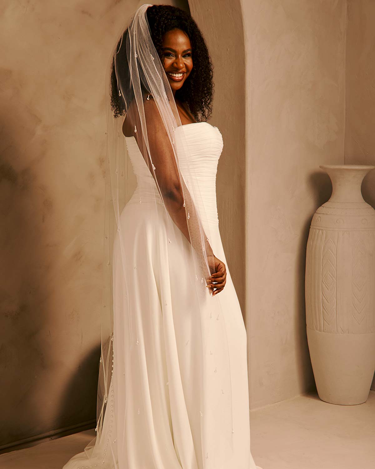 Bridal Veil with Crystal Raindrop Scatter Cassandra Lynne-A woman wearing a strapless ivory wedding gown with a long sheer veil, standing in a warm beige interior with a large decorative vase in the background.