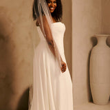 Bridal Veil with Crystal Raindrop Scatter Cassandra Lynne-A woman wearing a strapless ivory wedding gown with a long sheer veil, standing in a warm beige interior with a large decorative vase in the background.