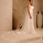 Rose Lace Royal Cathedral Bridal Veil Cassandra Lynne - A woman wearing a sleeveless wedding gown with a long sheer veil and lace-trimmed train, standing in a warm beige architectural setting with a large vase in the background.